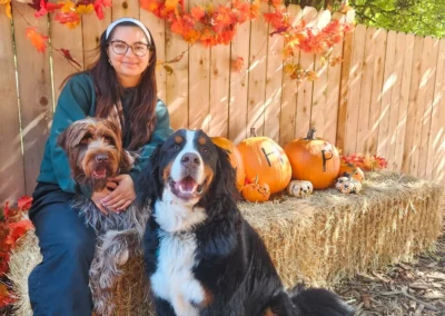 A camp counselor sits with two dogs on a hay bale in a fall photo setup with pumpkins at kennel-free boarding